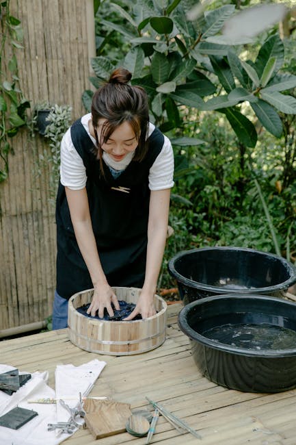 Asian woman standing outdoors tie-dyeing fabric, surrounded by greenery and natural elements.